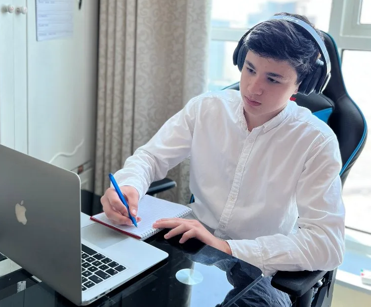 A pupil in headphones writing notes beside a laptop during a lesson at home